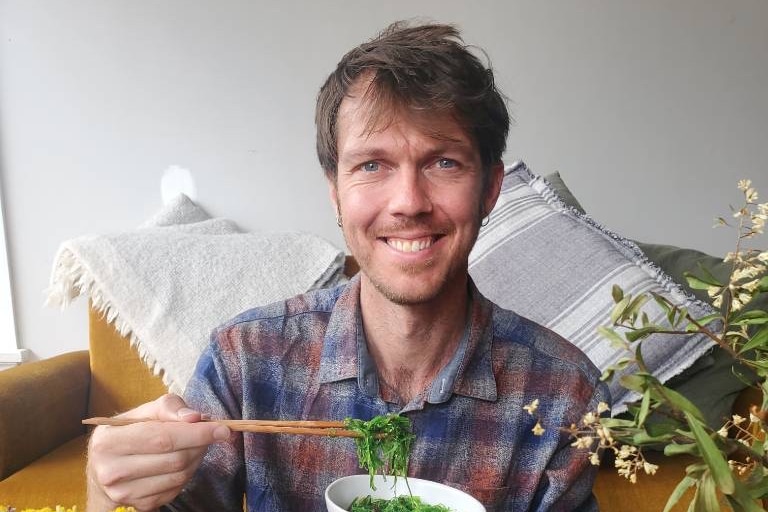 Man splies at camera holding chopsticks and a bowl of seaweed. 