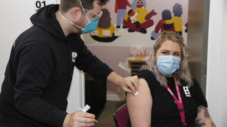 Woman in mask sitting on chair while man in mask holds vaccine near her arm.