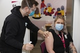 Woman in mask sitting on chair while man in mask holds vaccine near her arm.