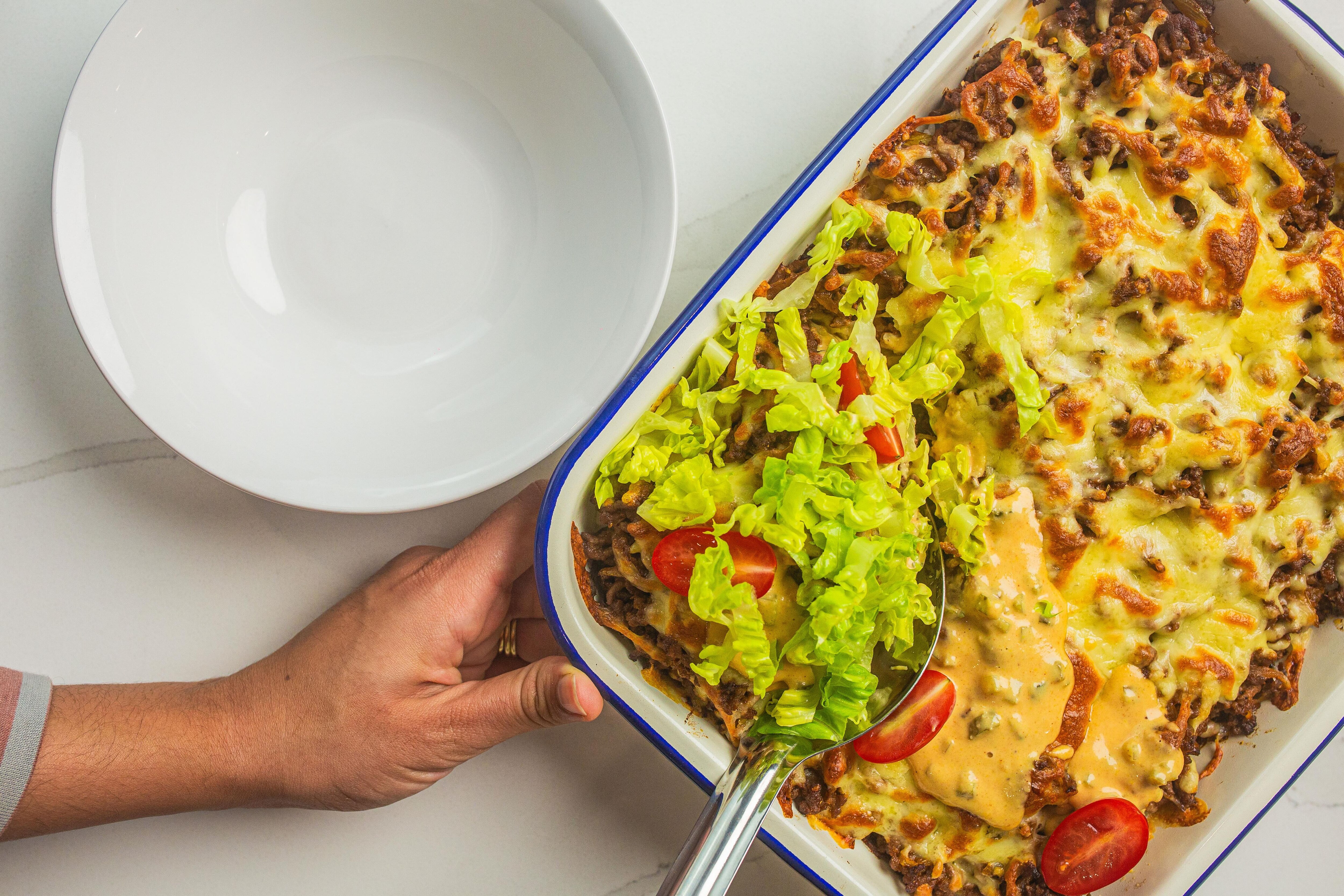 A woman scoops out a portion of cheeseburger rice bake from the baking tray to an awaiting bowl. A fun, delicious weeknight m