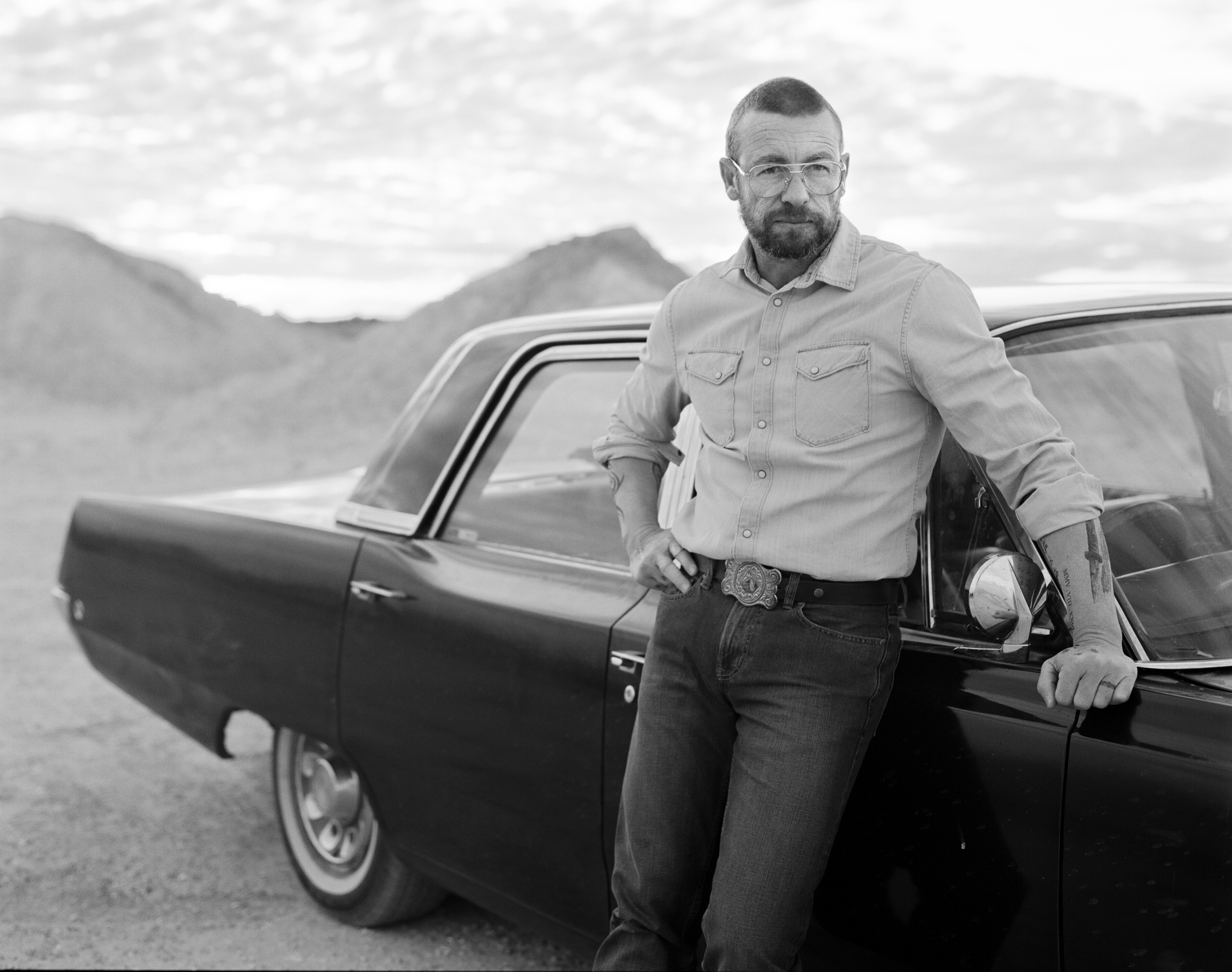 A black and white photo of a man with short hair and glasses leaning against an old car, in an outback Australian setting.