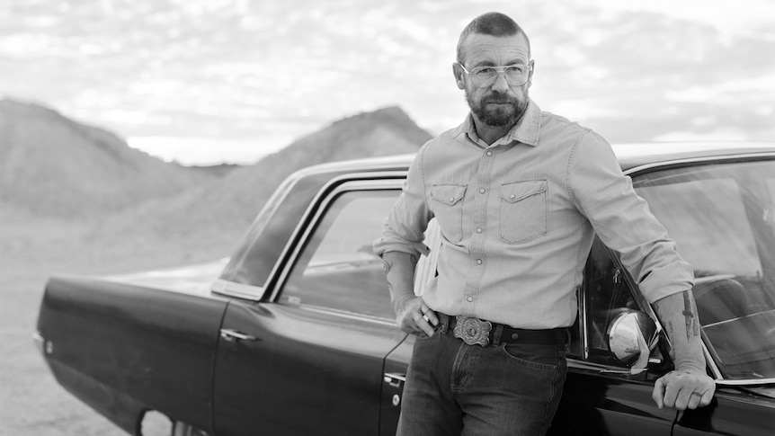 A black and white photo of a man with short hair and glasses leaning against an old car, in an outback Australian setting.