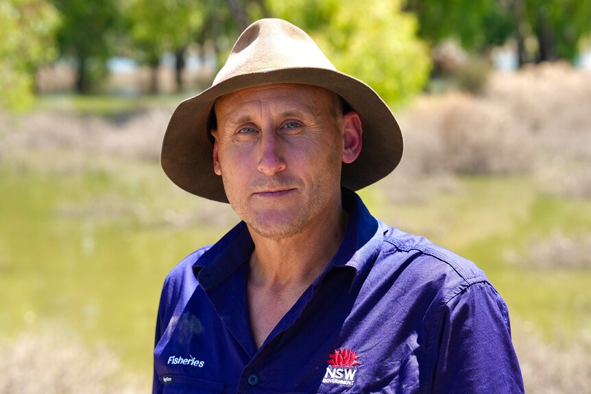 A white man with a blue shirt and brown hat standing in front of the Darling River.