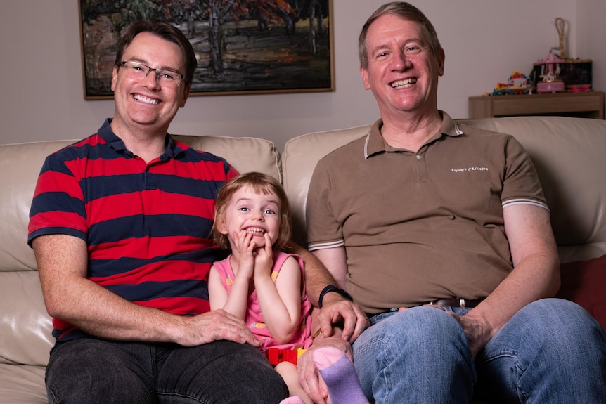 Two dads and a small girl on a couch.