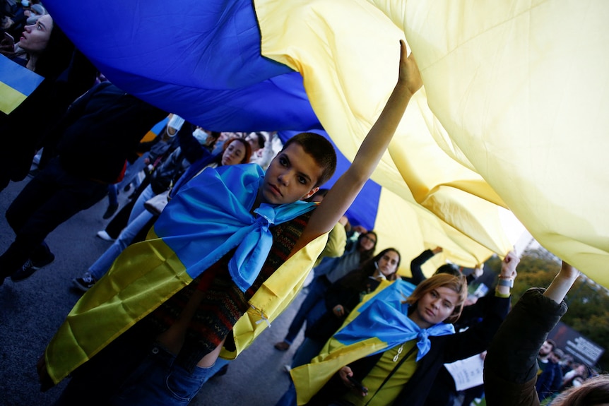 Protesters wrapped in Ukrainian flags hold up a larger flag with their hands while walking down a road.