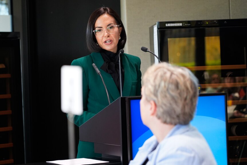 Prue Car wearing clear glasses and a green blazer at a podium in front of a microphone