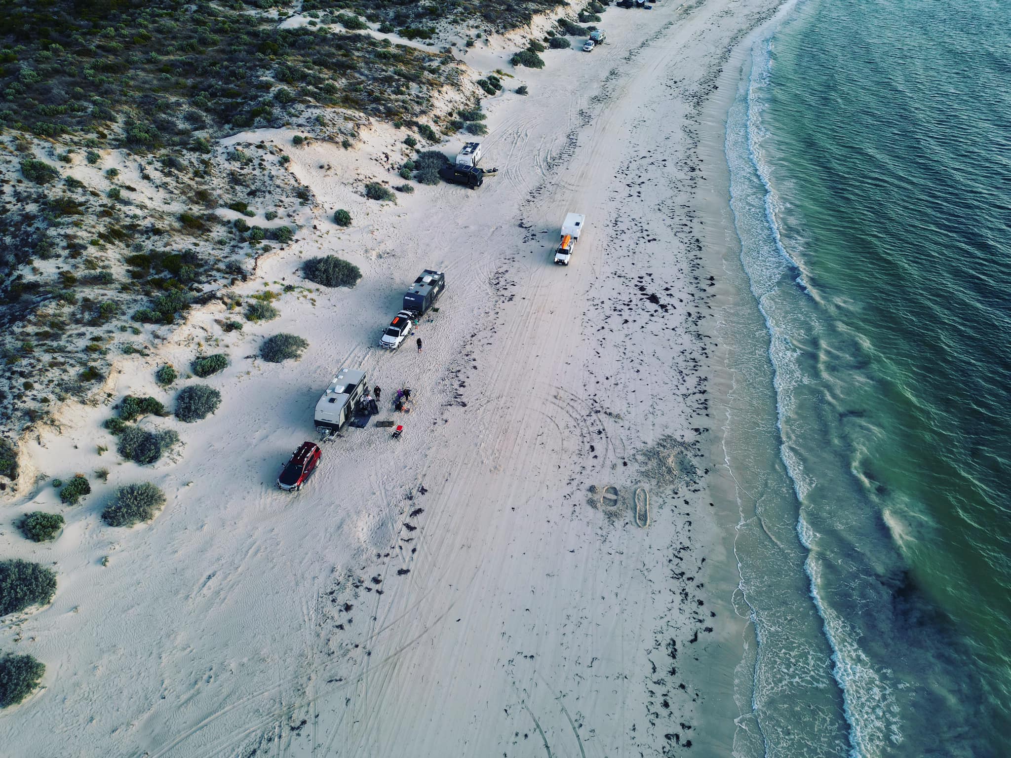 view from above of large vehicles camping on a beach with green water to the right