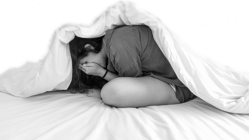 A young woman covers her eyes and cowers under a doona.