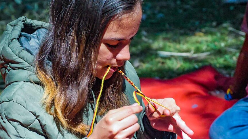 Girl weaves black, red and yellow yarn into a bracelet using her mouth and hands.