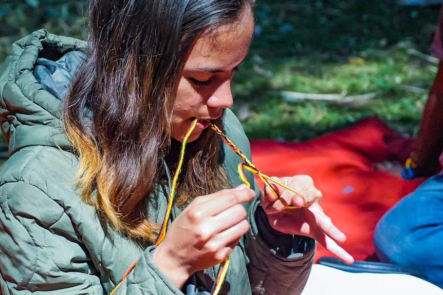 Girl weaves black, red and yellow yarn into a bracelet using her mouth and hands.