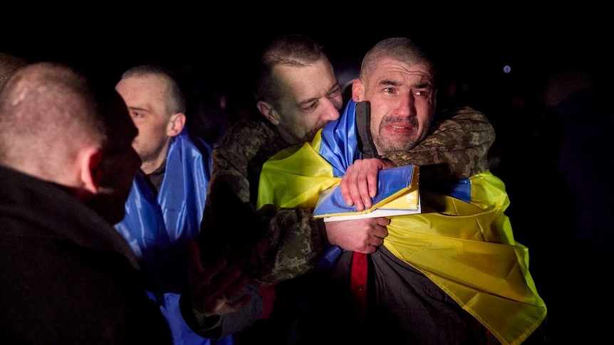 A man in combat fatigues hugs a man draped in the Ukranian flag