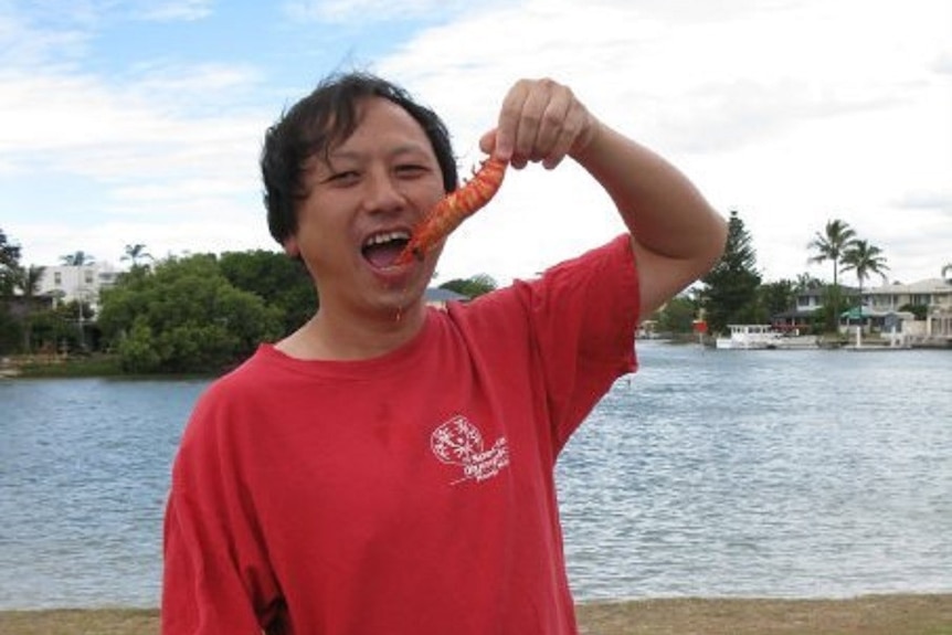 Man in red shirt smiles at camera while damgling a prawn near his open mouth.