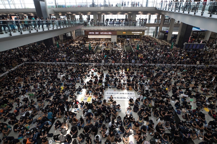 Hundreds of people sit on the floor an airport while on lookers watch from the upper level balconies