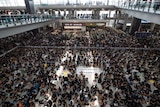 Hundreds of people sit on the floor an airport while on lookers watch from the upper level balconies