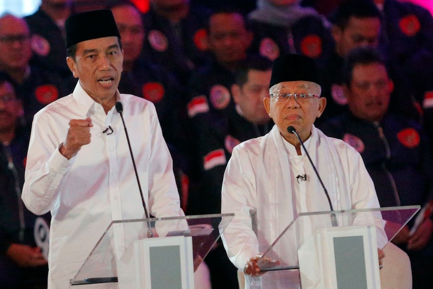 Two men standing at lecterns with microphones, one man gesturing with his fist as he speaks.