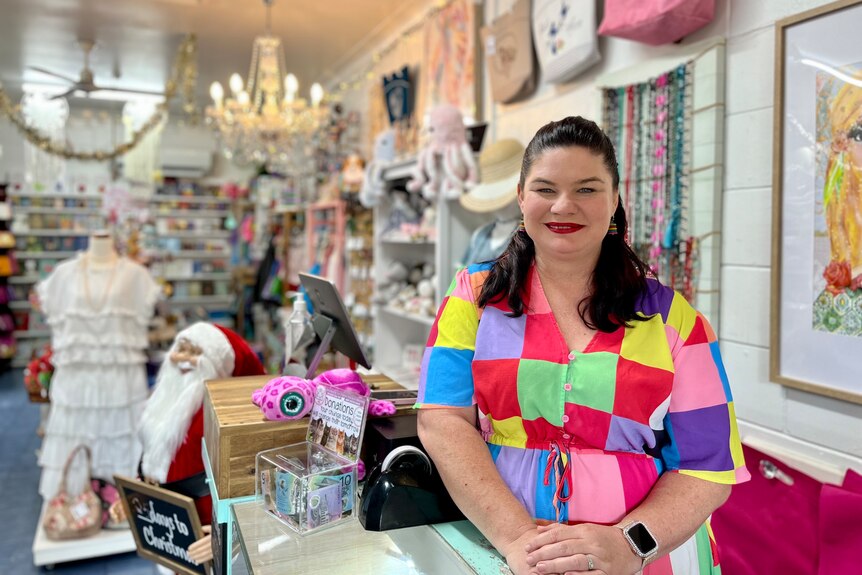 Mujer blanca con cabello largo y oscuro, con un vestido multicolor y de pie dentro de su tienda de regalos. 