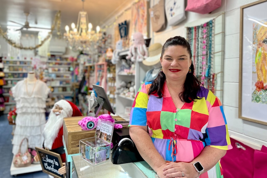 Femme blanche aux longs cheveux noirs, vêtue d'une robe multicolore et debout à l'intérieur de sa boutique de cadeaux. 