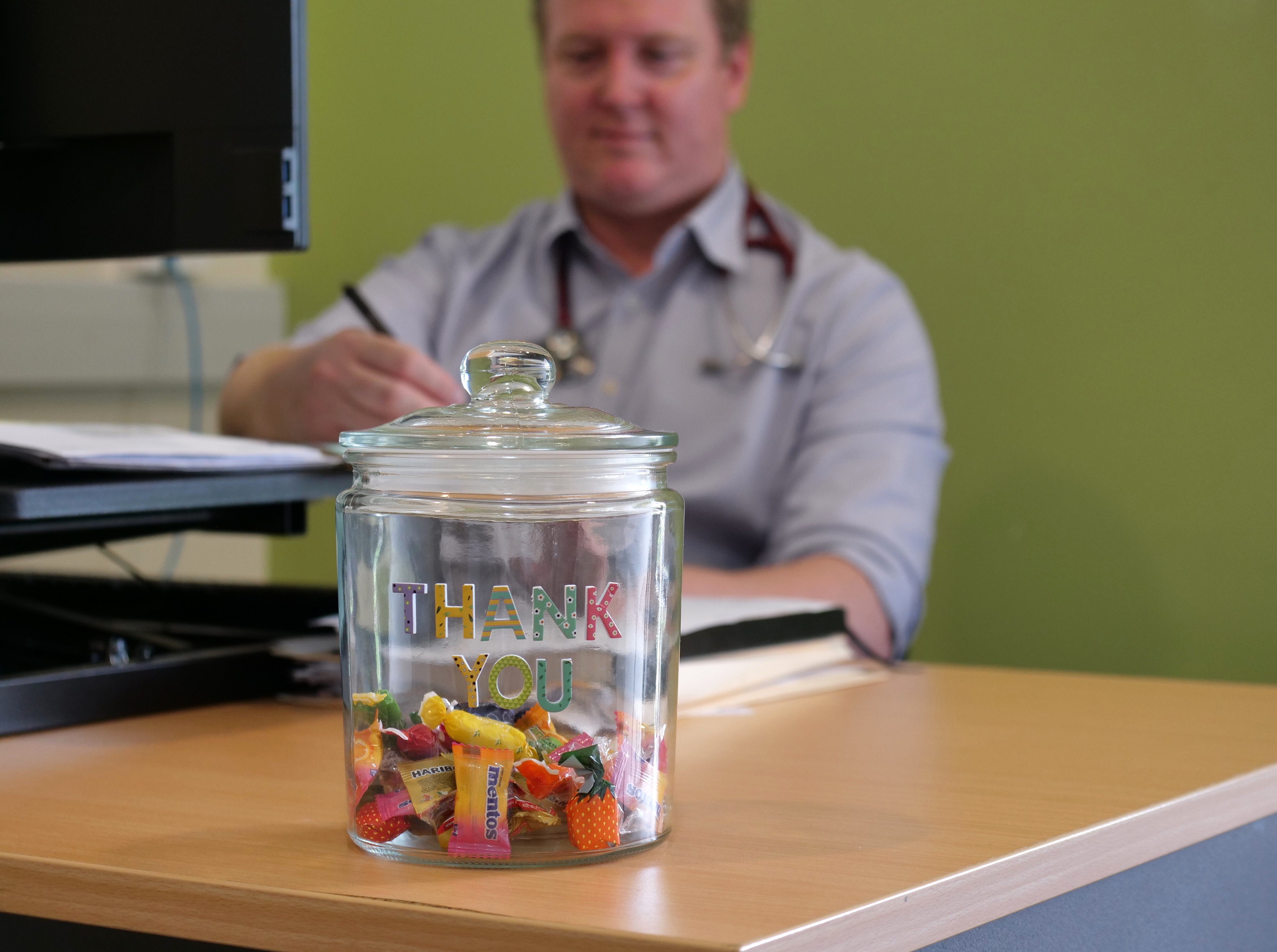 A doctor sits writing on his desk with a jar of lollies with thank you stickers on it