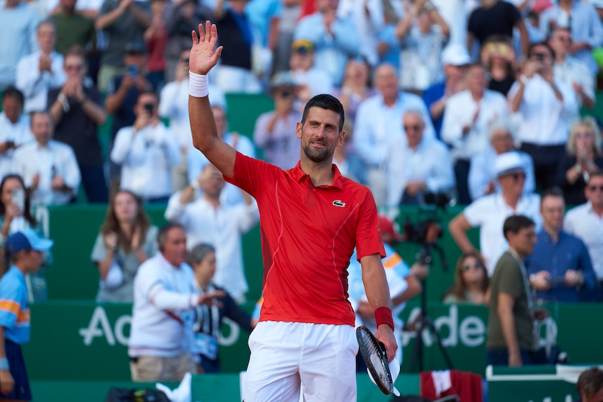 Tennis star Novak Djokovic stands on court and waves at the crowd after winning a match.