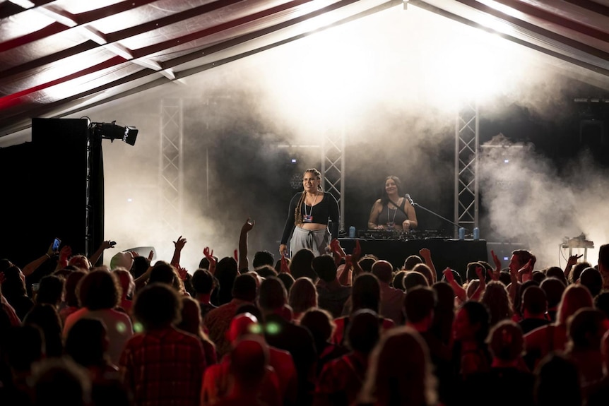 An Indigenous female rapper stands on a lit stage in front of a large crowd.