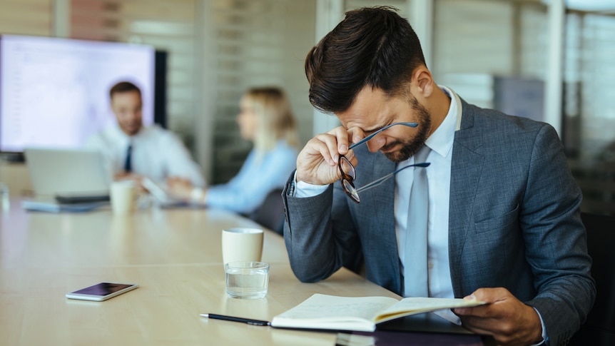 A tired young Caucasian man with brown hair squints his eyes to focus while holding glasses in hands in a meeting