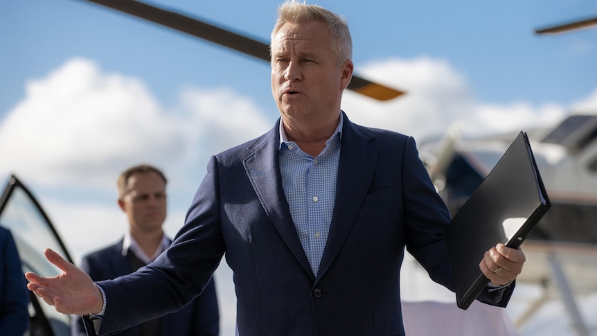 Jeremy Rockliff in a suit and holding a black folder gestures with both hands while standing in front of helicopters.