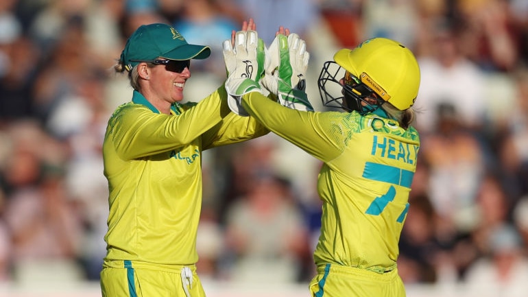 beth mooney and megan schutt give each other a double high five on the cricket pitch wearing gold