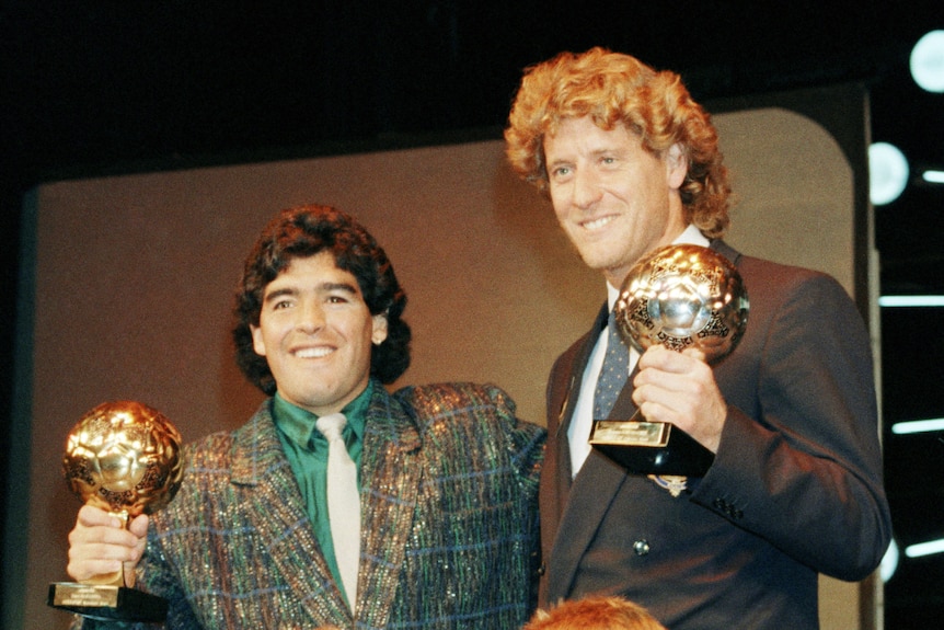 Two footballers posing with their gold trophies, with children by their side.