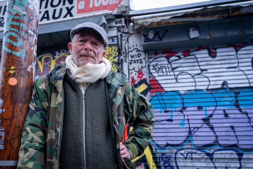 A man in a camo jacket and flat cap leans against a graffitied lamp post.
