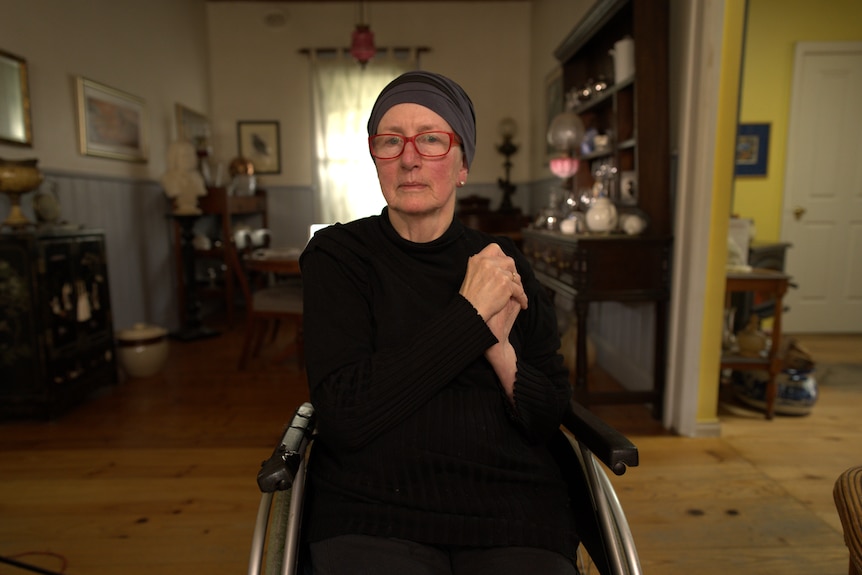 Woman wearing a black top and pants, sitting in a wheelchair with her hands clasped.