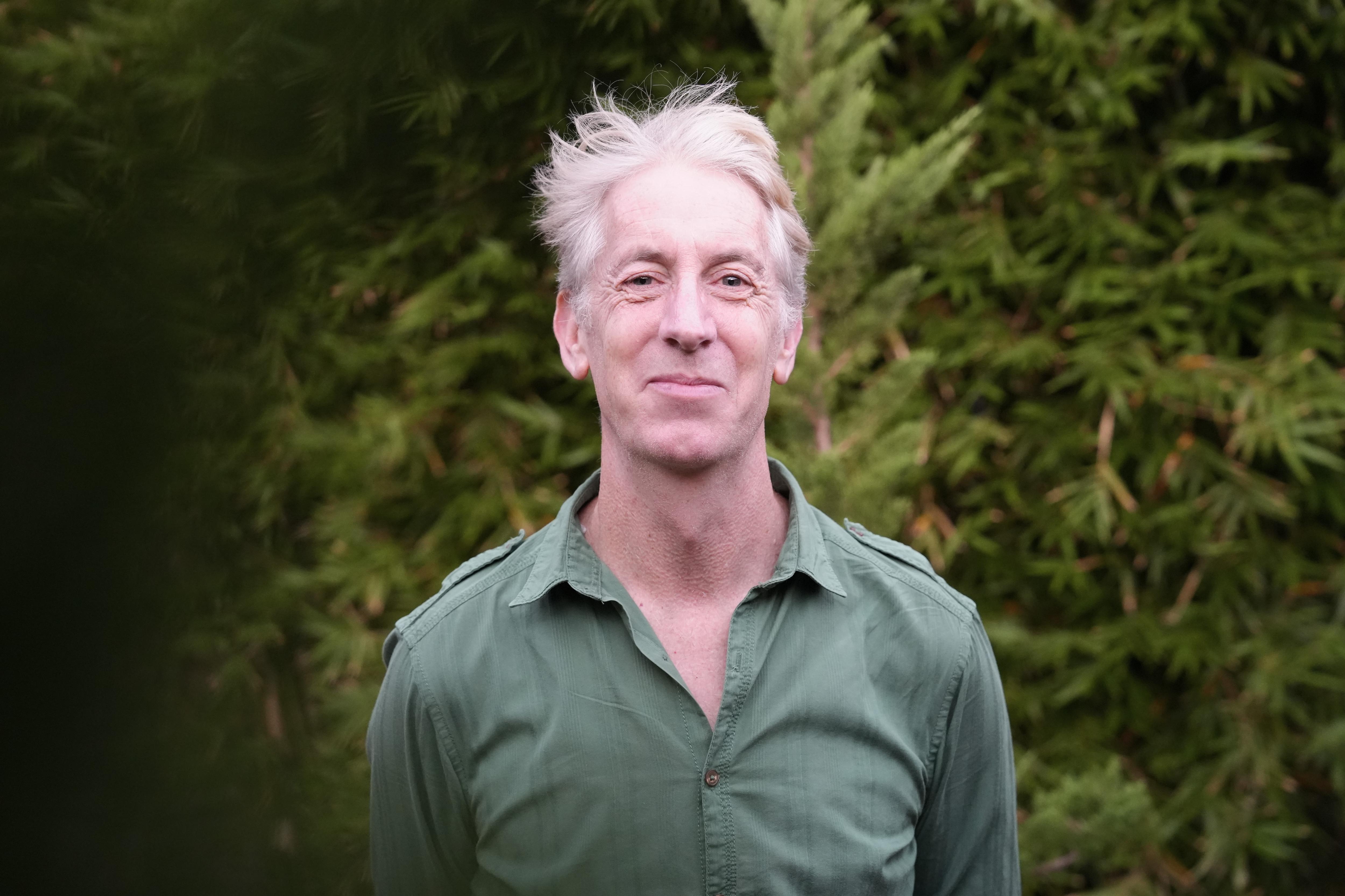 Un hombre con cabello gris y una camisa verde frente a un árbol