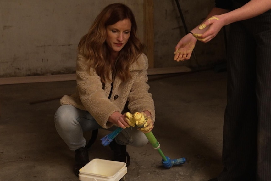 A woman crouches over a tub of paint holding a plastic object. 