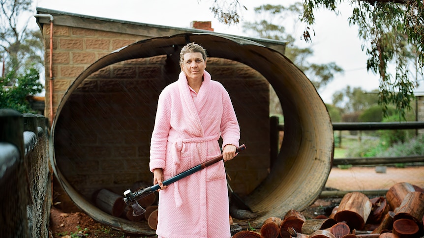 Claudias mother lisa stands in front of firewood with an axe in hand wearing pink pajamas
