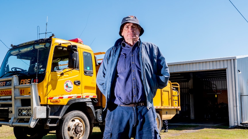 A man in a rural fire uniform stands in front of a fire truck. He is missing his left arm.