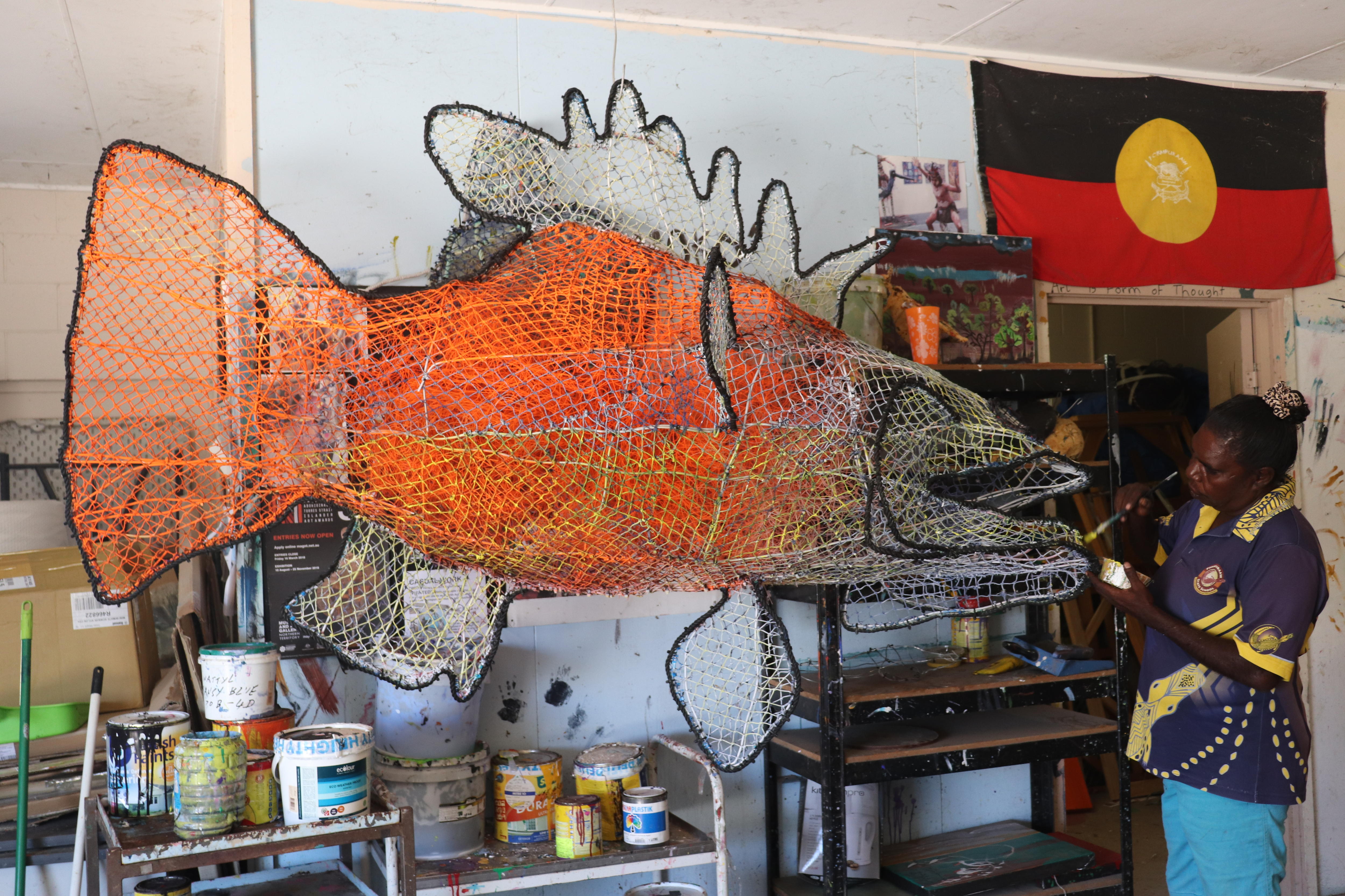 A First Nations man woman stands in an art studio painting a large fish sculpture made from netting.