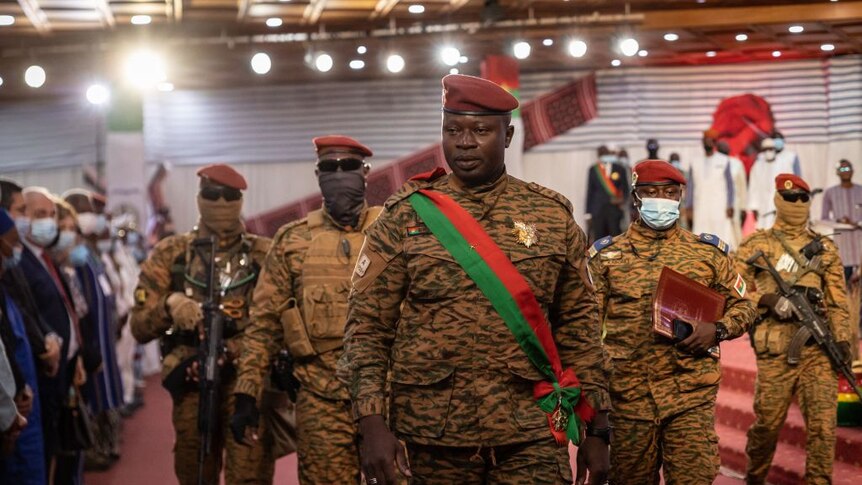 An African man in military camouflage uniform wearing a red and green sash and a maroon beret walks a red carpet