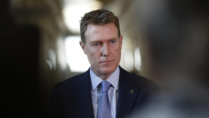 A man with brown hair wearing a suit and tie looks to the right, he is in focus with people closer to the camera out of focus 