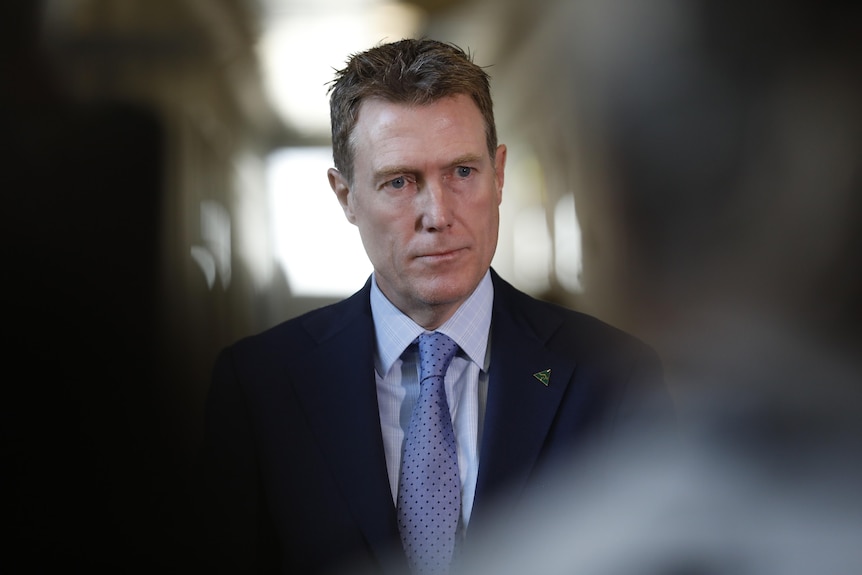 A man with brown hair wearing a suit and tie looks to the right, he is in focus with people closer to the camera out of focus 