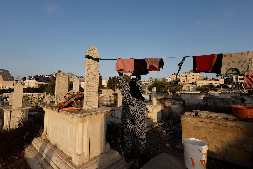 A woman in a headscarf hangs laundry on a line strung between two grave stones. 