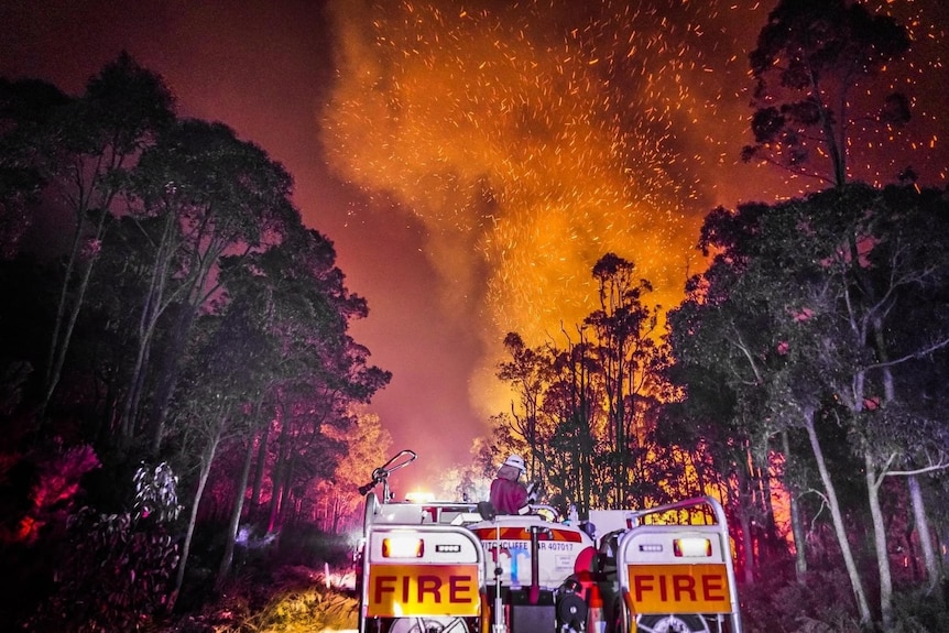 A fire truck in the foreground with flames and embers leaping into the night sky in front of them.