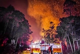 A fire truck in the foreground with flames and embers leaping into the night sky in front of them.