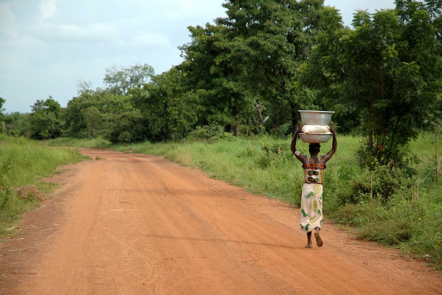 A woman carries water on her head along a dirt road, with grass and large trees in the background.