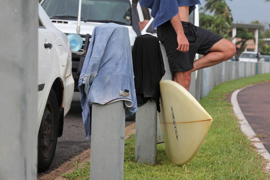 A surfboard, some t-shirts and a surfer leaning against posts on a street near the beach.