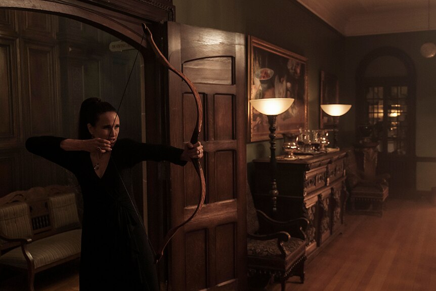 Andie MacDowell stands with focused expression in dimly lit Gothic style room with drawn bow.