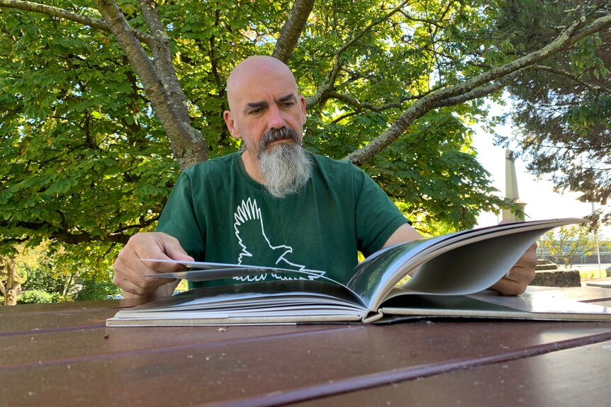A man sits at a picnic table under a tree and looks through a book of photographs