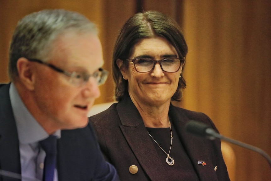 Michelle Bullock sits at a table and smiles as Philip Lowe, sitting next to her, speaks into a microphone.