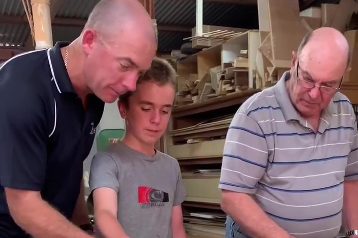 Dad on left, son in middle, grandfather on right, Dad guiding son with sprayer putting glue on board in workshop