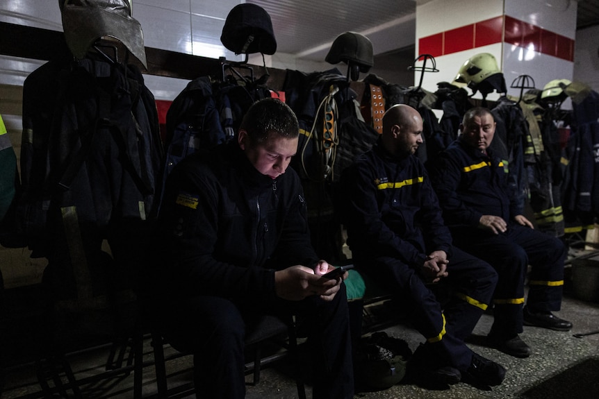 a group of firefighters sit on a bench inside a fire station with their firefighting gear behind them