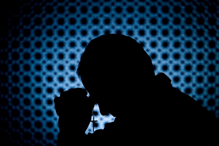 Silhouette of priest bowing head and holding cross, against blue and black background.
