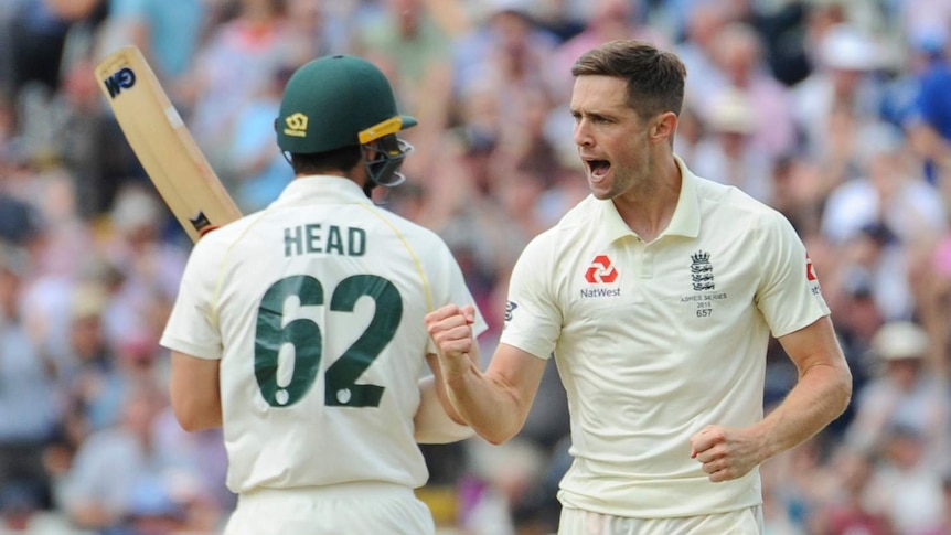 England bowler Chris Woakes pumps his fist as he runs past Australia batsman Travis Head, seen from behind.